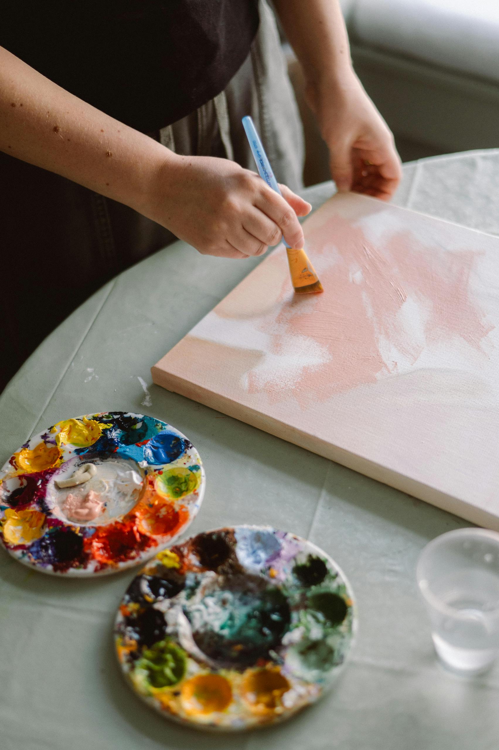A female artist works on an abstract painting with vibrant colors on a sunlit studio table.